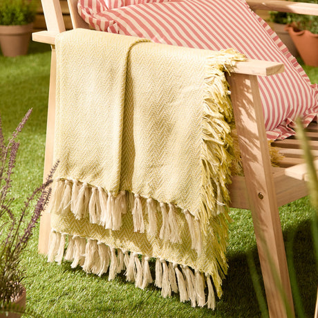 Wisley Herringbone Polyester Throw in yellow and cream draped over a wooden chair arm with long fringed tassels, shown outdoors on grass beside striped cushions.