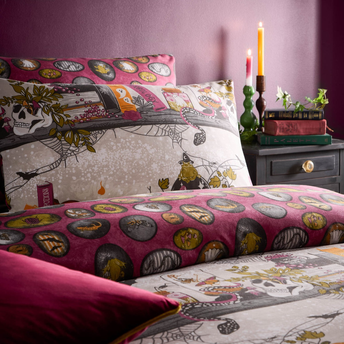 Reversible duvet cover set in a multicolor Halloween print with skulls, spell books and cobwebs on one side and a pink oval pattern on the reverse, shown on a bed in a pink-toned bedroom with candles.