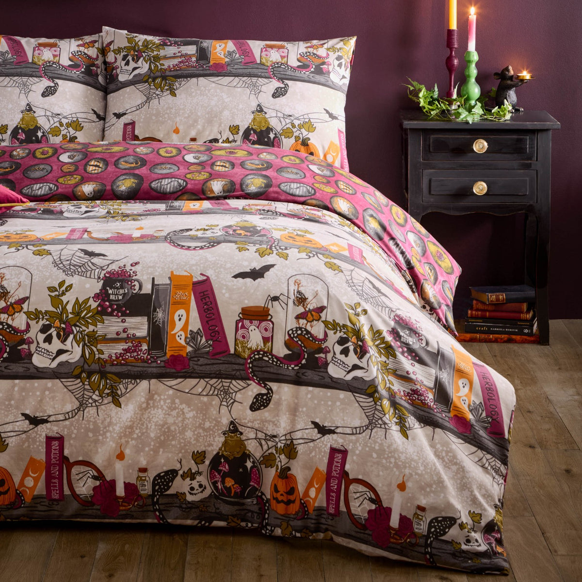 Reversible Witchy Tales Duvet Cover Set on a bed, cream Halloween side with skulls, pumpkins, spell books and jars, deep pink reverse, in a dark candlelit bedroom.