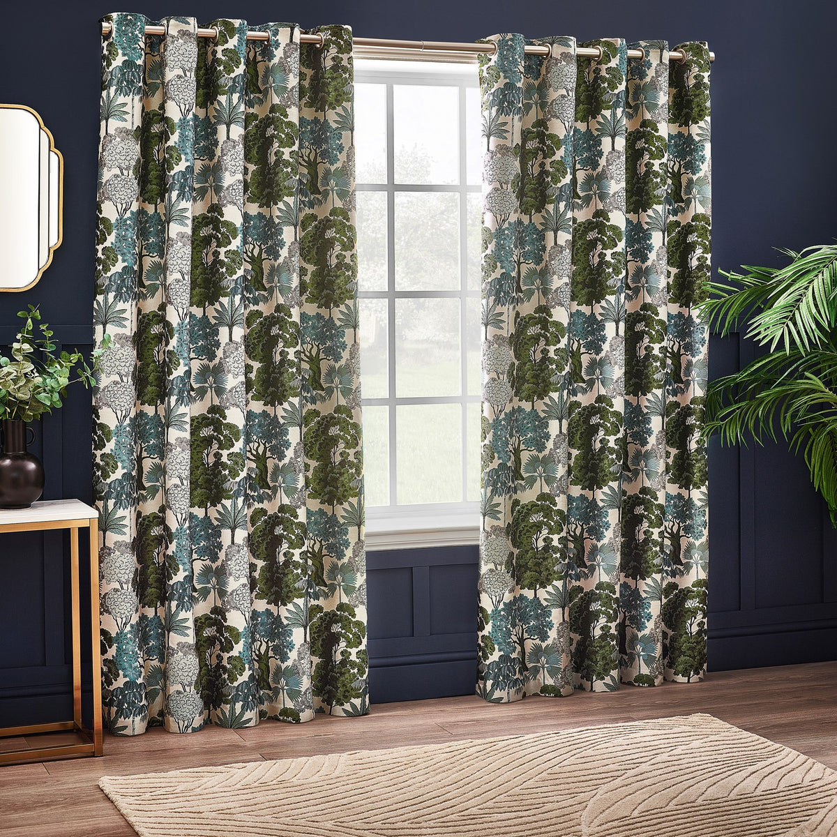 Woodlands Jacquard fully lined eyelet curtains in green, showing a woven woodland tree pattern in green, teal and grey on cream, hung by a window in a navy-walled living room.