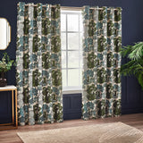 Woodlands Jacquard fully lined eyelet curtains in green, showing a woven woodland tree pattern in green, teal and grey on cream, hung by a window in a navy-walled living room.