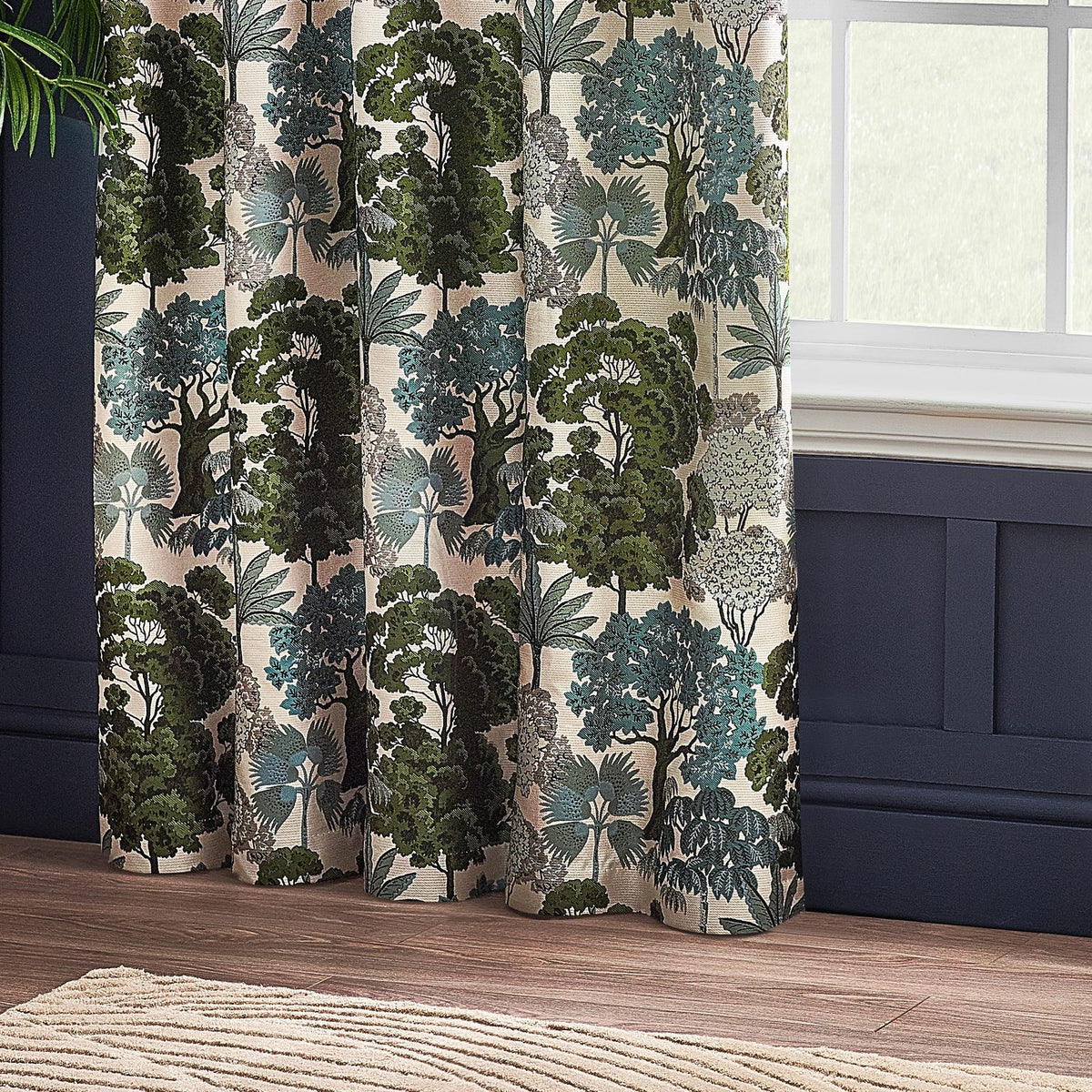 Woodlands Jacquard Fully Lined Eyelet Curtains in green, showing a woven woodland scene of trees and palms on cream fabric, hung by a window in a room with navy panelling.