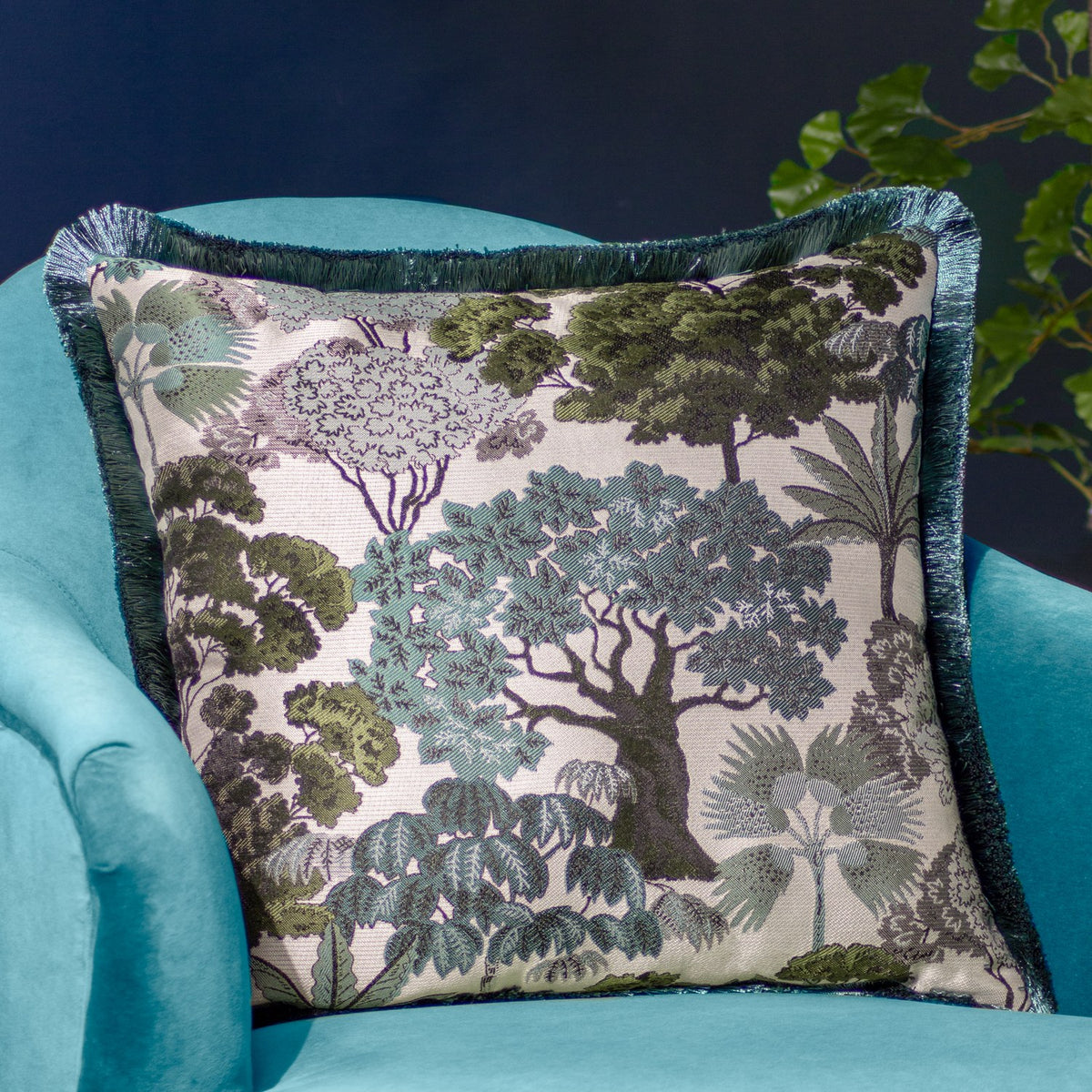 Woodlands Eyelet Velvet Cushion in green, square jacquard design with woven tree canopy pattern in sage and grey tones, teal fringe trim, shown on a turquoise velvet armchair.