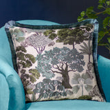 Woodlands Eyelet Velvet Cushion in green, square jacquard design with woven tree canopy pattern in sage and grey tones, teal fringe trim, shown on a turquoise velvet armchair.