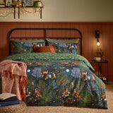 Woodland Ritual Printed Reversible Duvet Cover Set in navy on a bed, showing a moonlit forest scene with deer, foxes and owls, paired with green pillowcases and a rust throw in a bedroom setting.