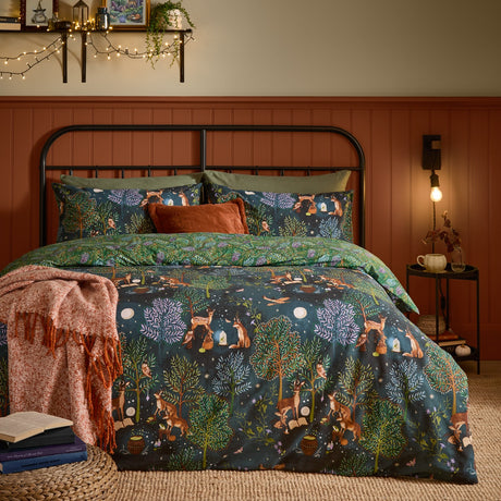 Woodland Ritual Printed Reversible Duvet Cover Set in navy on a bed, showing a moonlit forest scene with deer, foxes and owls, paired with green pillowcases and a rust throw in a bedroom setting.