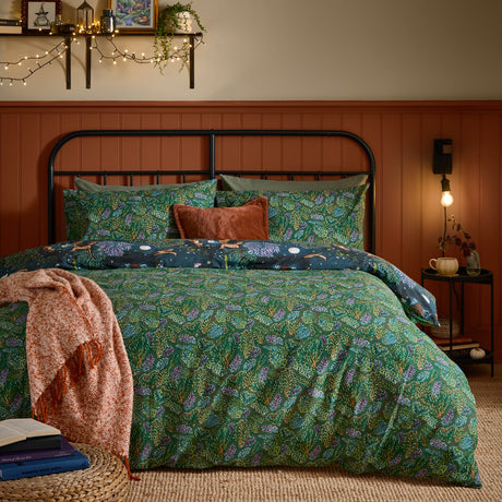 Woodland Ritual reversible duvet cover set on a bed, showing the green foliage side with navy woodland-print pillowcases, styled in a bedroom with a rust-toned throw.
