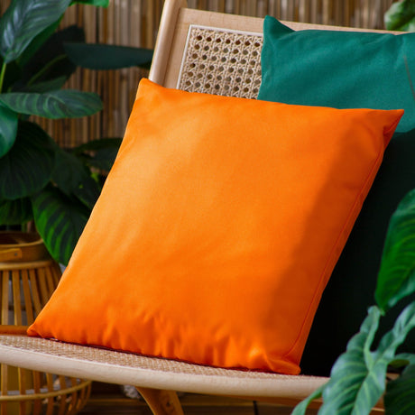 Square orange polyester cushion with a knife-edge finish, shown on a rattan chair with a green cushion behind it and green foliage around.