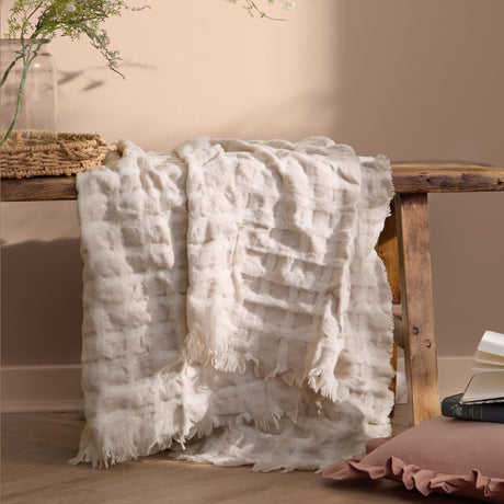 Walton Waffle Check Linen Throw in cream draped over a wooden bench, showing its waffle-weave grid texture and fringed edge in a warm-toned room setting.