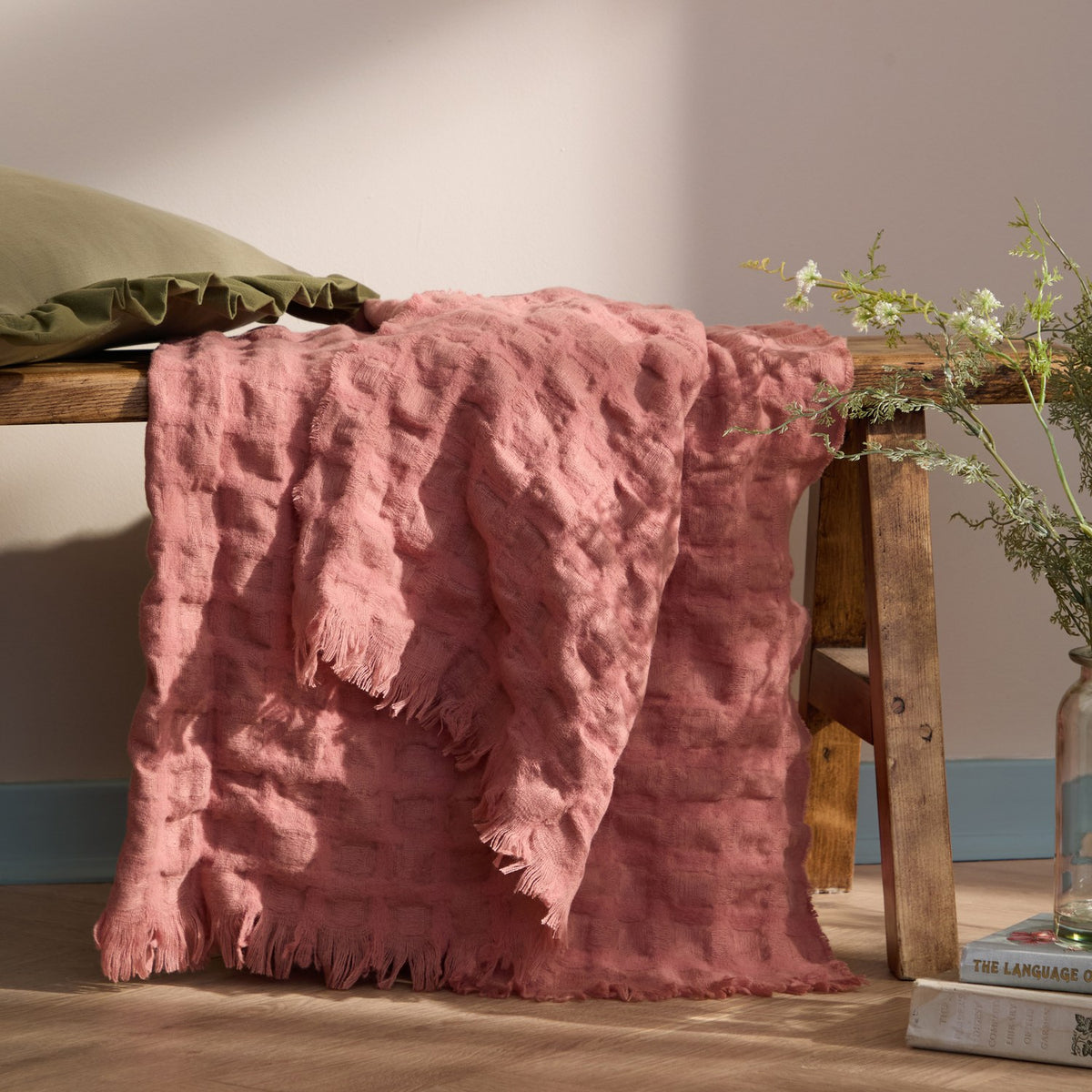 Walton Waffle Check Polyester Throw in rose pink with raised waffle-textured grid and fringed edges, draped over a wooden bench in a living room setting.