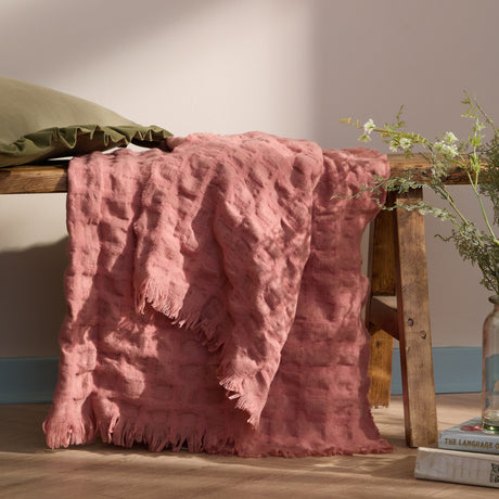 Walton Waffle Check Polyester Throw in rose pink with raised waffle-textured grid and fringed edges, draped over a wooden bench in a living room setting.
