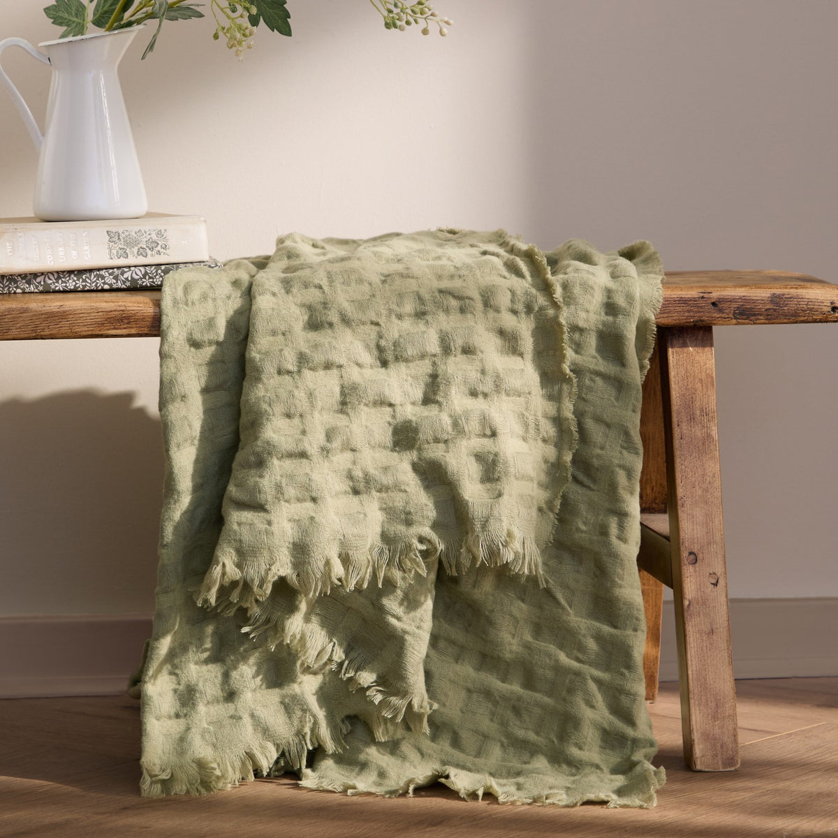 Walton Waffle Check Polyester Throw in sage green with fringed edges, draped over a wooden bench beside a jug of foliage and books.