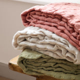 Folded Walton Waffle Check Polyester Throw in sage green with fringed edge, stacked with pink and cream throws on a wooden stool.