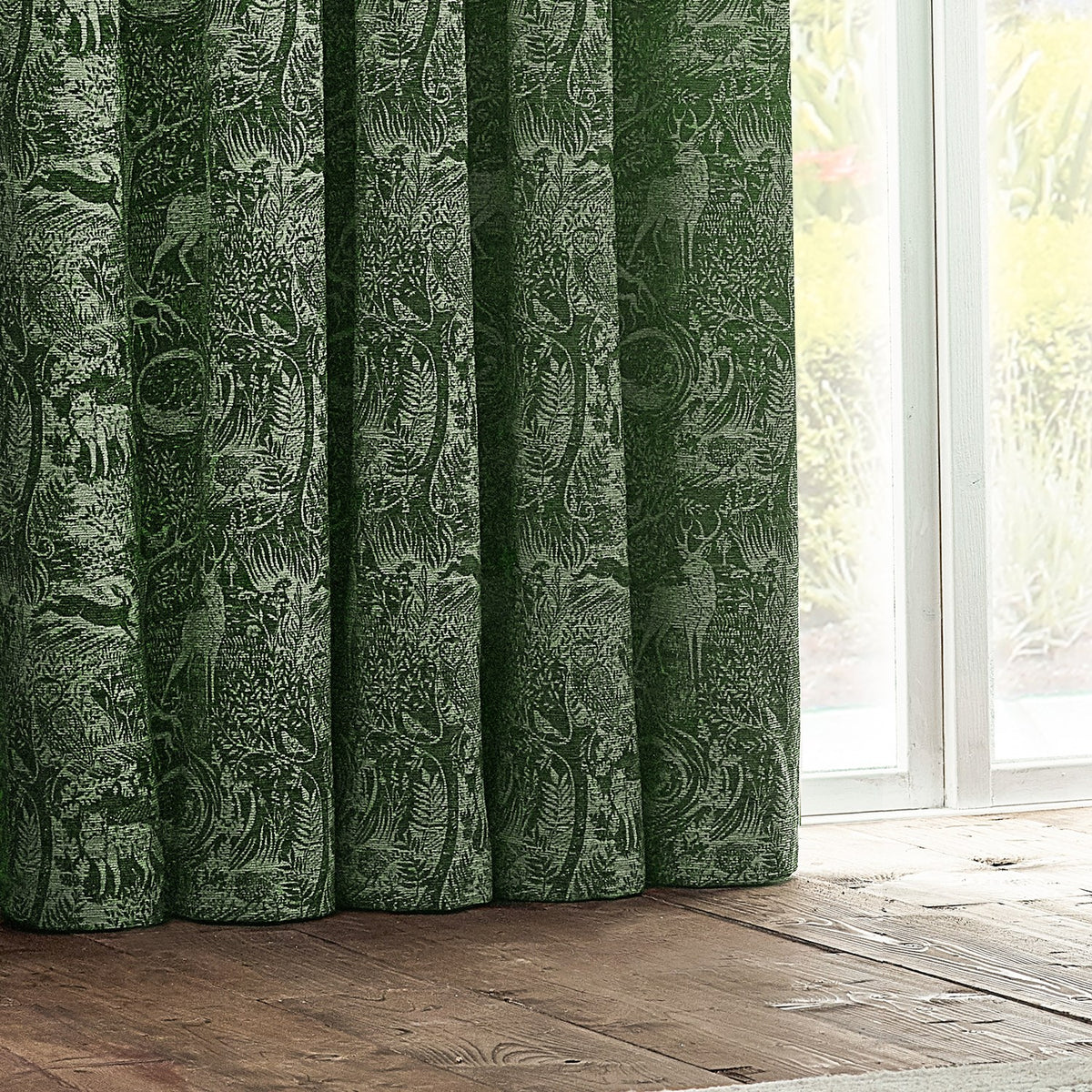 Winter Woods Chenille Lined Eyelet Curtains in emerald, showing folded panels with a tonal woodland motif of deer and birds, hung beside a window in a room setting.