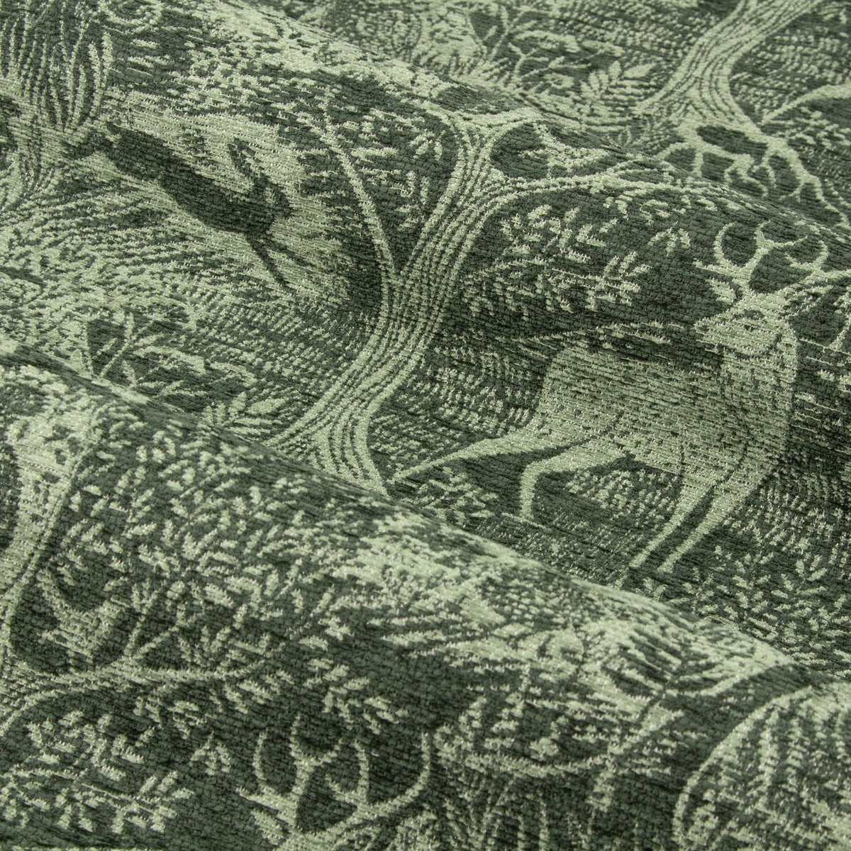 Close-up of Winter Woods eyelet curtain fabric in emerald, showing a chenille jacquard woodland design with deer, hares, birds and trees woven in tonal green.