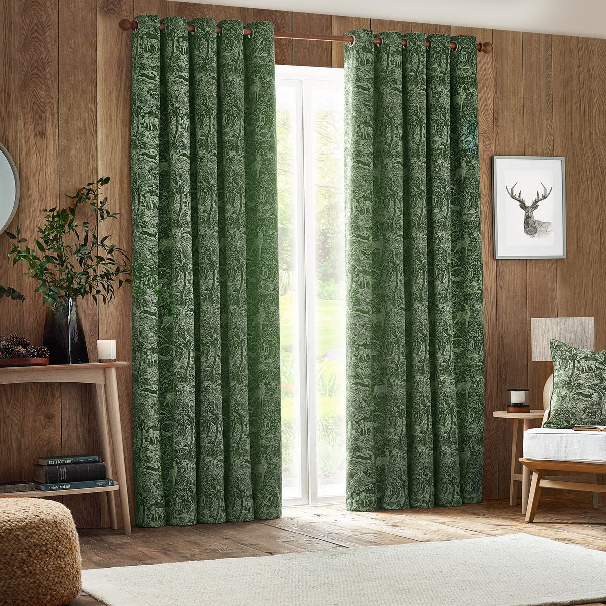 Winter Woods Chenille Lined Eyelet Curtains in emerald, shown floor-length beside a glass door in a wood-panelled living room with a woodland jacquard pattern.