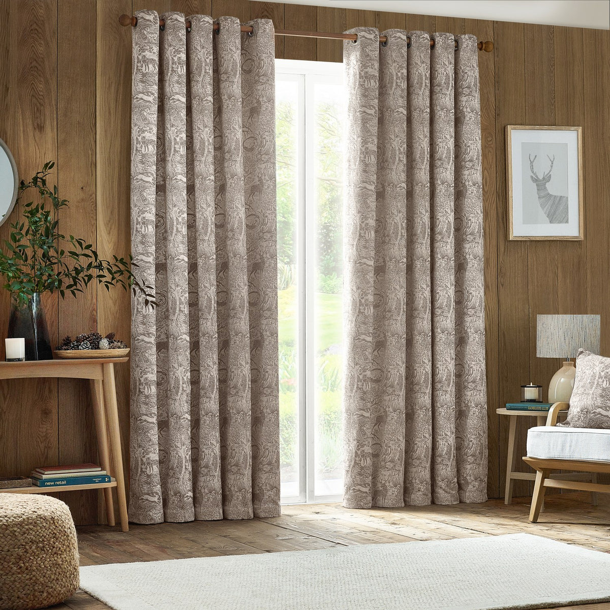 Winter Woods Chenille Lined Eyelet Curtains in taupe with woodland animal jacquard weave, hung on a wooden pole beside a glass door in a wood-panelled living room.