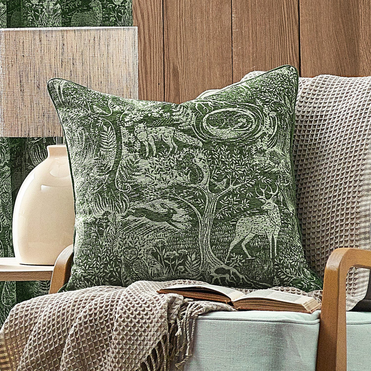 Winter Woods Jacquard Chenille Cushion in emerald, woven with a woodland scene of deer, hare, squirrel and trees, shown on an armchair with a beige waffle throw.