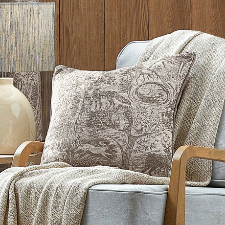 Winter Woods Jacquard Chenille Cushion in taupe with woven woodland animal and tree design, shown on a light blue armchair beside a neutral throw.