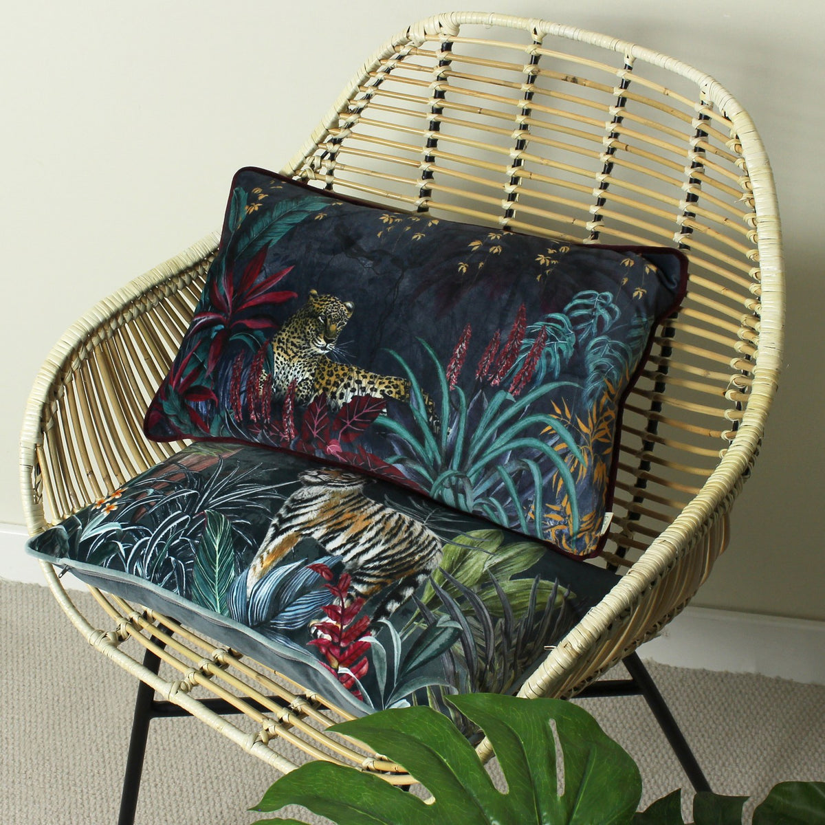 Zinara Animal Velvet Cushion in black with hand-painted leopard and jungle foliage design, shown on a rattan chair above a tiger-print cushion.