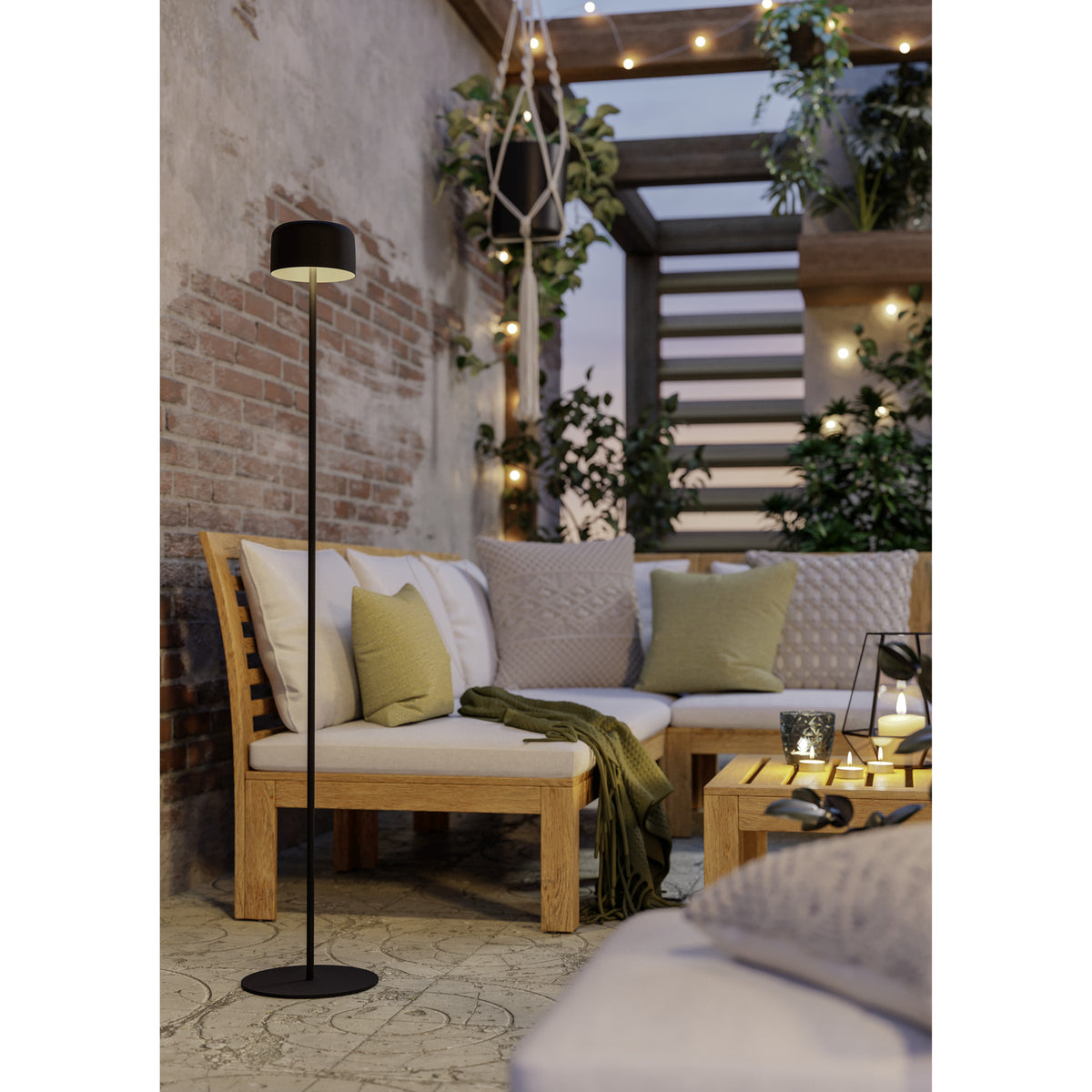 Roccanova-L solar LED floor lamp in black steel with a drum-shaped head and round disc base, shown lit on an outdoor patio beside a sofa.