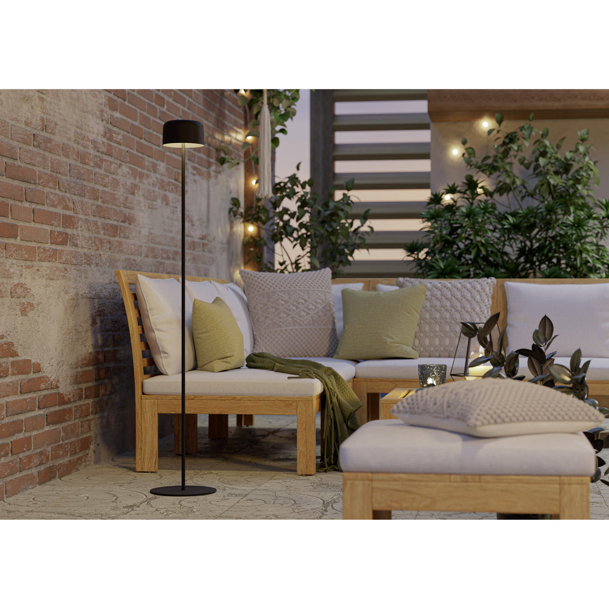 Roccanova-L solar LED floor lamp in black steel with a slim stem, drum-shaped head and round disc base, shown beside an outdoor corner sofa with cushions.