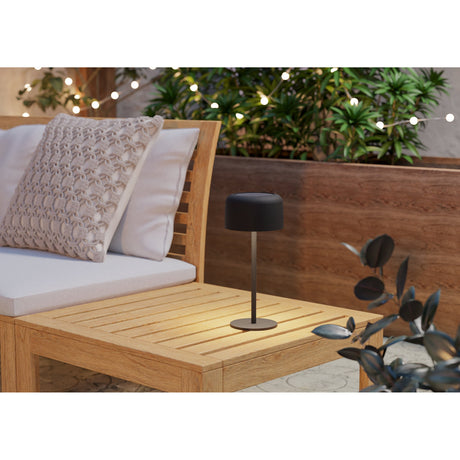 Roccanova solar LED touch table lamp in black steel with domed shade and slim stem, lit on an outdoor wooden side table beside a sofa.