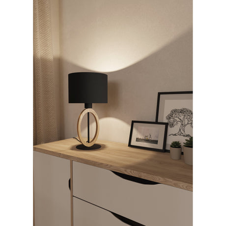 Rolmanne Table Lamp 42.5cm in black and brown, featuring a black fabric drum shade and oval wood ring on a round black steel base, shown on a wooden sideboard in a bedroom setting.
