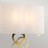 The Rosa Large 2 Light Wall Light - Aged Brass showcases a modern and elegant design with interconnected aged brass rings supporting an ivory faux silk rectangular lamp shade. This soft-white shade emits a gentle glow, enhancing the overall ambiance.