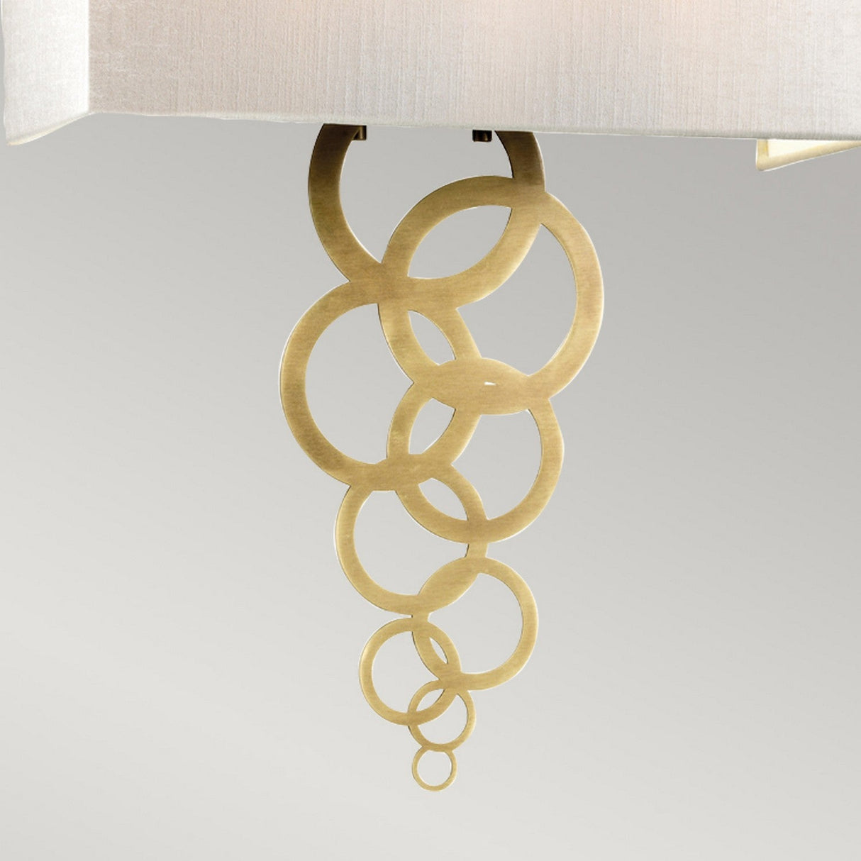 The Rosa Large 2 Light Wall Light in Aged Brass showcases a modern and elegant design with interconnected, aged brass rings of varying sizes cascading from its base, beautifully complemented by an ivory faux silk shade set against a plain gray background.