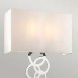 The Rosa Large 2 Light Wall Light in polished chrome features a rectangular fabric shade that emits a warm glow, complemented by a decorative metal element with interlocking circles, which adds an elegant touch to its contemporary design.