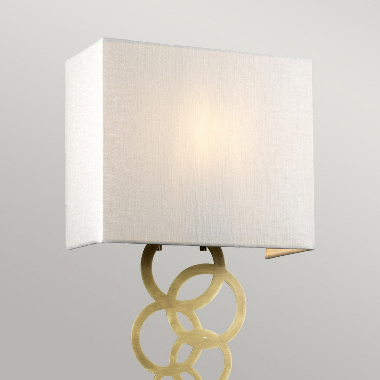 The Rosa Medium 1 Light Wall Light in aged brass features a square, ivory faux silk shade and a decorative gold metal base with interlocking circular rings. Its vintage charm softly illuminates the space, casting a warm and inviting glow.