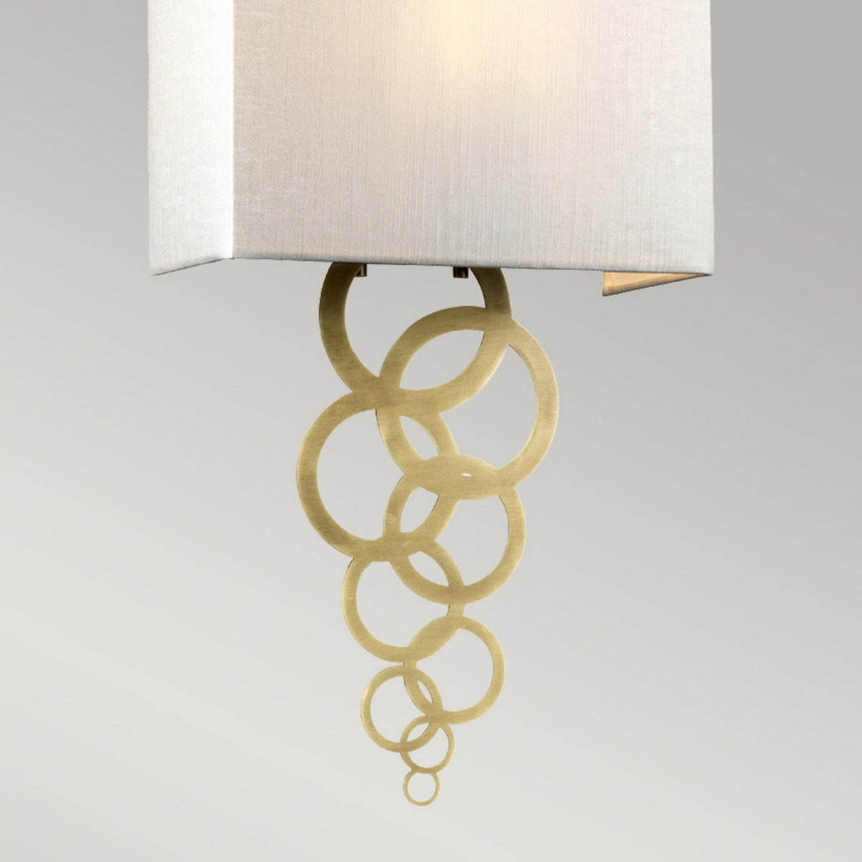 The Rosa Medium 1 Light Wall Light in Aged Brass features a modern design with a rectangular ivory faux silk shade, complemented by decorative gold rings cascading below, exuding vintage charm against a neutral background.