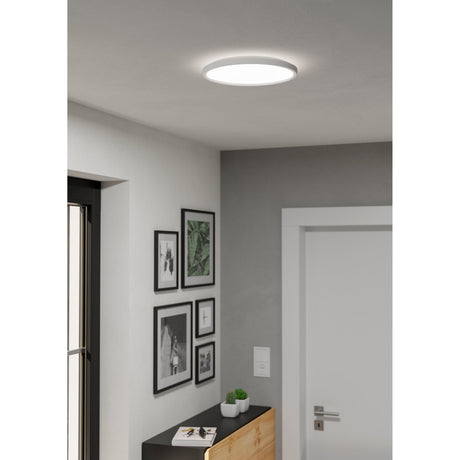 Rovito 30cm round LED flush ceiling light in white plastic, illuminated on a hallway ceiling above a gallery wall of framed pictures.