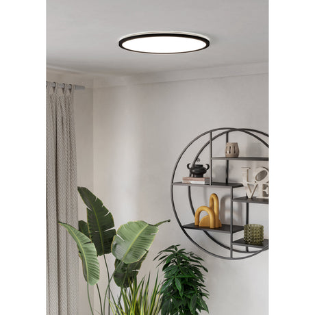 Rovito 50cm LED flush ceiling light with black rim and white diffuser, shown illuminated on a white ceiling in a living room with a wall shelf and plants.