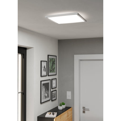 Rovito square LED ceiling panel light in white plastic, 39 x 39cm, shown illuminated on a hallway ceiling above framed artwork and a door.