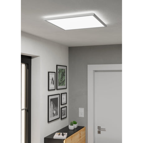Rovito square LED ceiling panel light, 50 x 50cm, white plastic frame with smooth diffuser, installed flush on a grey hallway ceiling above a gallery wall of framed photos.