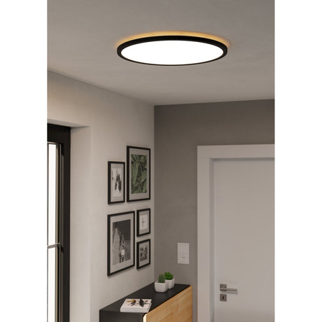Rovito 50cm round LED flush ceiling light with black frame, white diffuser and warm backlit edge, shown installed in a hallway with gallery frames below.