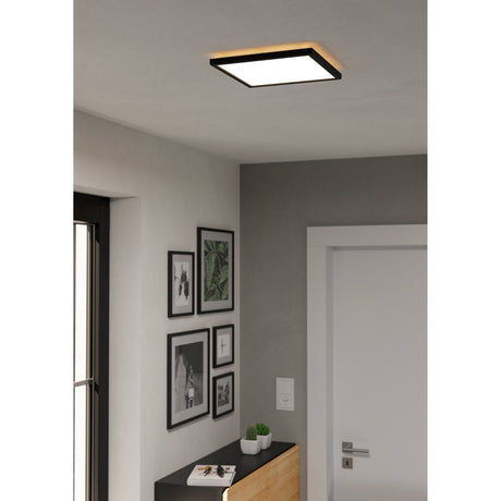 Rovito Smart LED Panel Light, square black-framed ceiling fixture with white diffuser and warm glow, shown mounted in a hallway with grey walls, framed pictures and a white door.