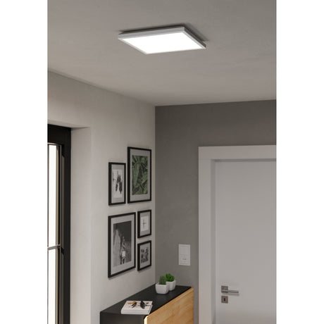 Rovito Smart Colour Changing LED Panel Light, 29 x 29cm square white ceiling fixture, shown recessed into a grey hallway ceiling above a gallery wall and wooden sideboard.