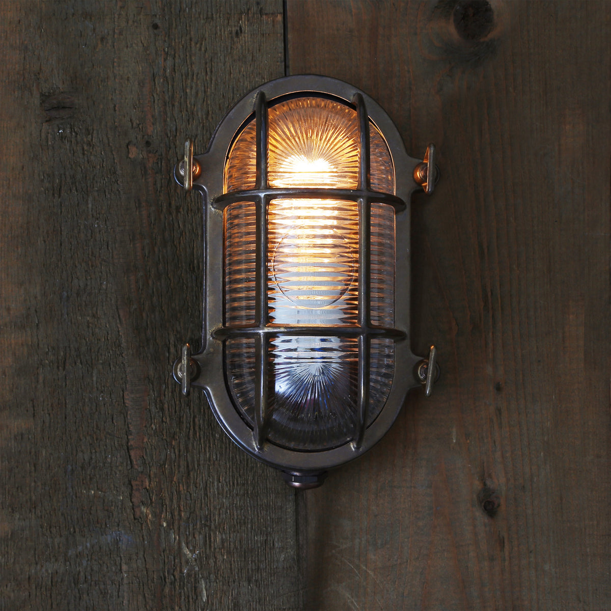 Ruben Small Bulkhead wall light in brass with a caged oval body over ribbed glass, lit on a dark wooden wall, IP54 rated.
