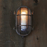 Ruben Small Bulkhead wall light in brass with a caged oval body over ribbed glass, lit on a dark wooden wall, IP54 rated.