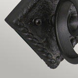 Close-up of the Rectory 1 Light Small Wall Light in black, featuring a twisted rope design around its edges and set against a textured square base. Its thick, curved structure complements rustic interiors, evoking a touch of medieval decor, while the neutral gray background highlights its charm.