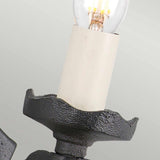 A detailed look at the Rectory 1 Light Wall Light in black showcases its candle-style bulb holder with an exposed light bulb. The wrought iron fixture boasts a textured finish and an uneven edge design that mimics a melted candle, adding to its rustic charm. This medieval-inspired lighting stands out against a simple light gray background.