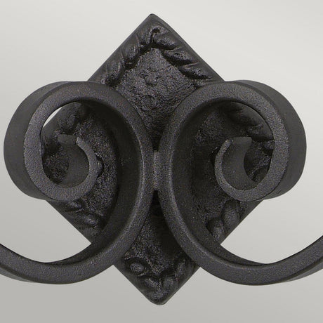 Close-up of the Rectory 2 Light Small Wall Light in black, showcasing an intricate wrought iron scrollwork design with twin spirals on both sides against a simple light gray background. The centerpiece highlights a diamond-shaped element adorned with elaborate details, evoking a medieval-inspired aesthetic.