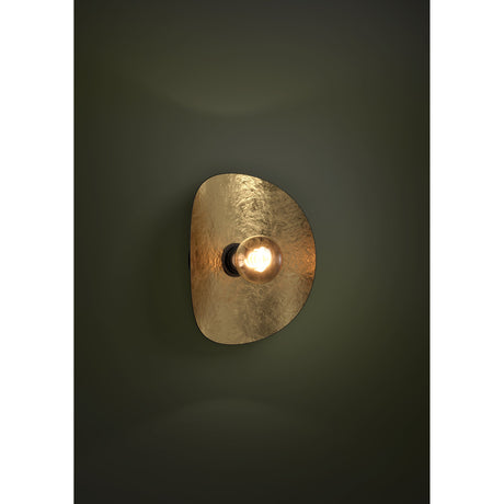 Saceda wall light in black and gold, an irregular curved steel shade with a hammered gold interior and black exterior housing a lit E27 bulb, mounted on a dark green wall.