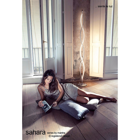 A woman in a light dress lounges on cushions, reading near tall shuttered windows. Behind her, the Sahara LED Floor Lamp 21W 3000K in Silver & Chrome casts gentle ambient lighting across the wooden floor.
