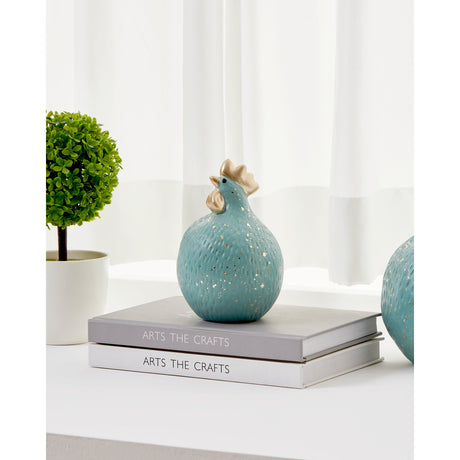 Saidane ceramic cockerel ornament in muted mint green with gold comb, wattles and speckled texture, displayed on stacked books beside a potted plant.