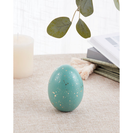 Saidane ceramic decorative egg in dark green with gold-flecked matt finish, displayed upright on a fabric surface beside a candle, books, and foliage.