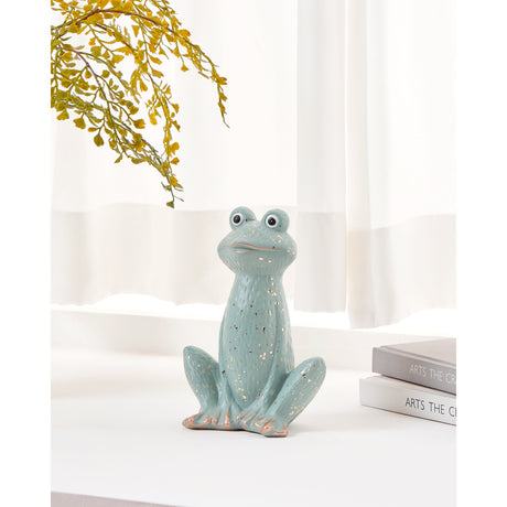 Saidane ceramic frog ornament in mint green with gold speckles, painted eyes and gold-tipped toes, seated on a table near books and a sheer curtain.